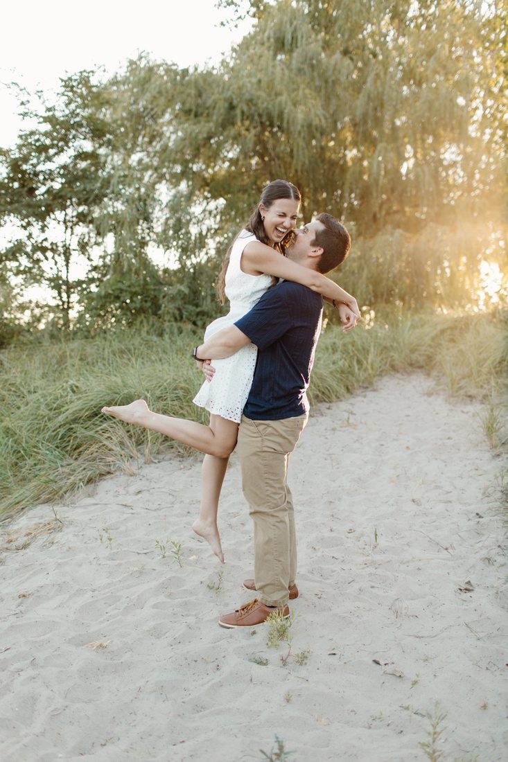 Couple embracing on a sandy dune path at golden hour, he lifts her as she laughs — Home Team Photography