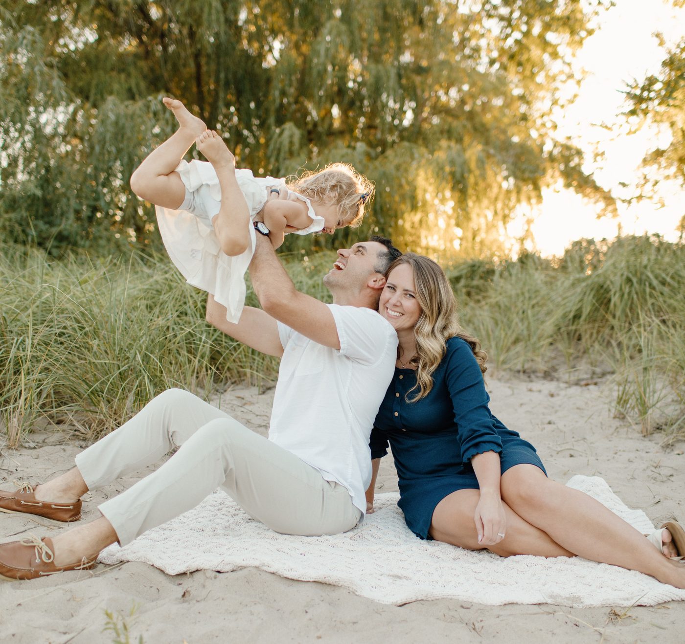 Parents lifting their laughing toddler on a beach blanket at golden hour — Home Team Photography