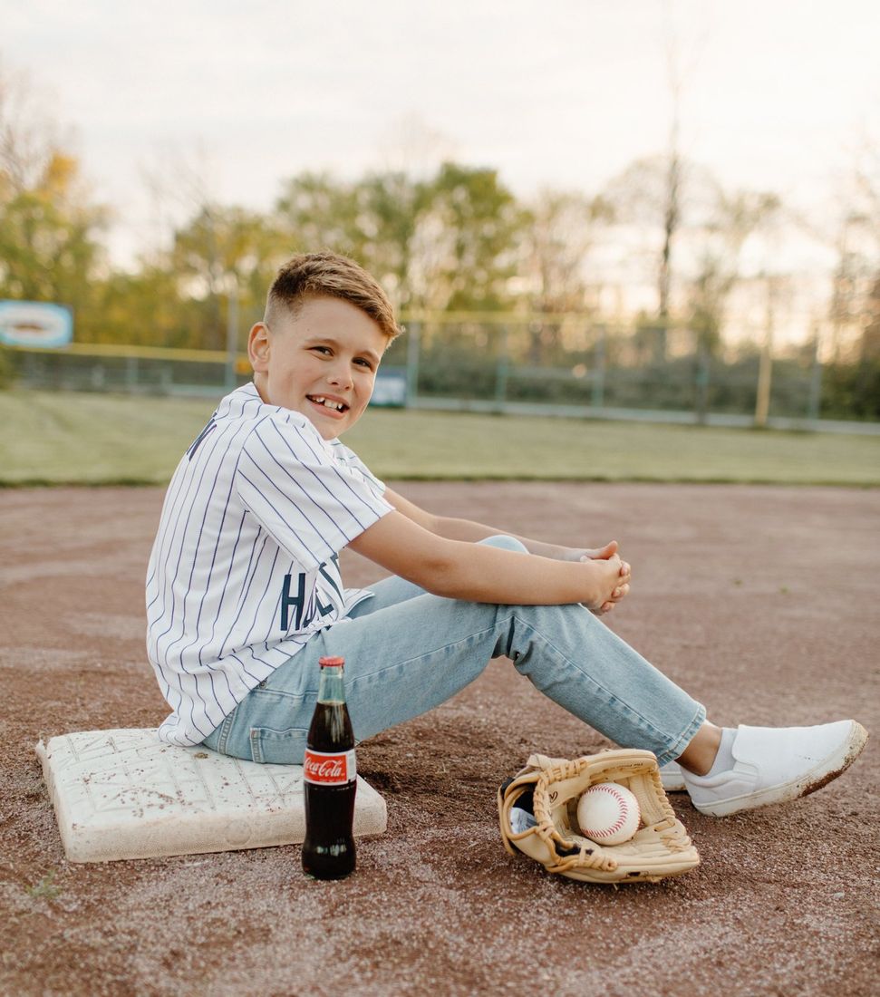 Boy in a pinstripe baseball jersey seated on the field at sunset — Home Team Photography