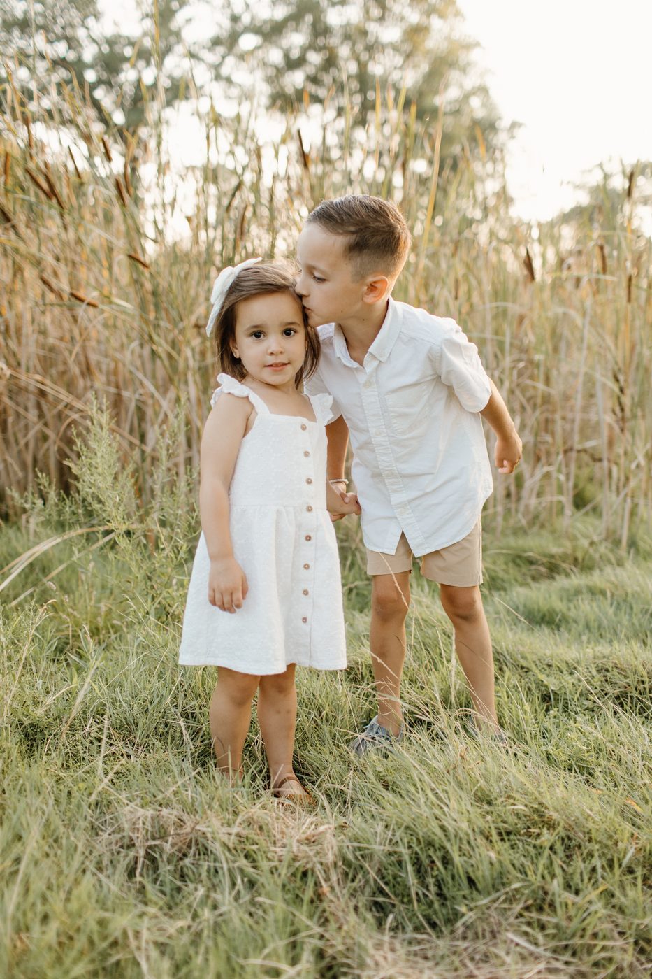 Big brother kissing his little sister on the head in tall grass — Home Team Photography