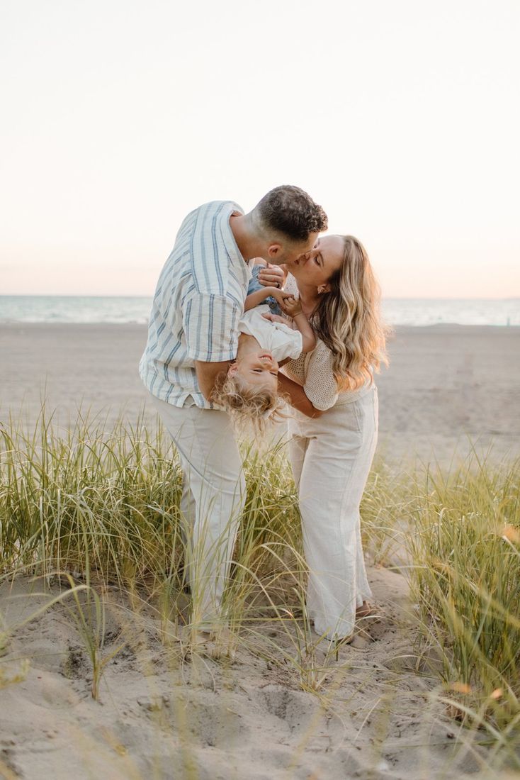 Family at golden hour on a Western New York beach — parents kissing while dad dips their laughing toddler — Home Team Photography