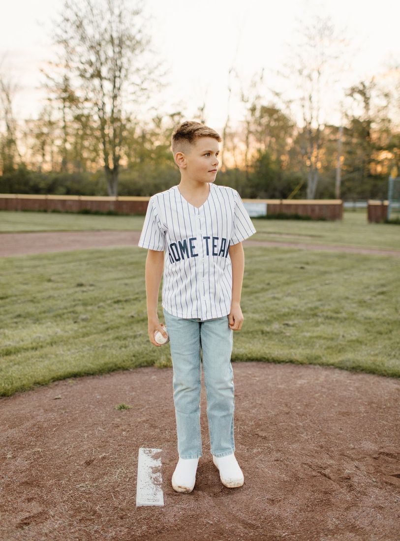 Young boy in a Home Team baseball jersey holding a ball on the field at golden hour — Home Team Photography