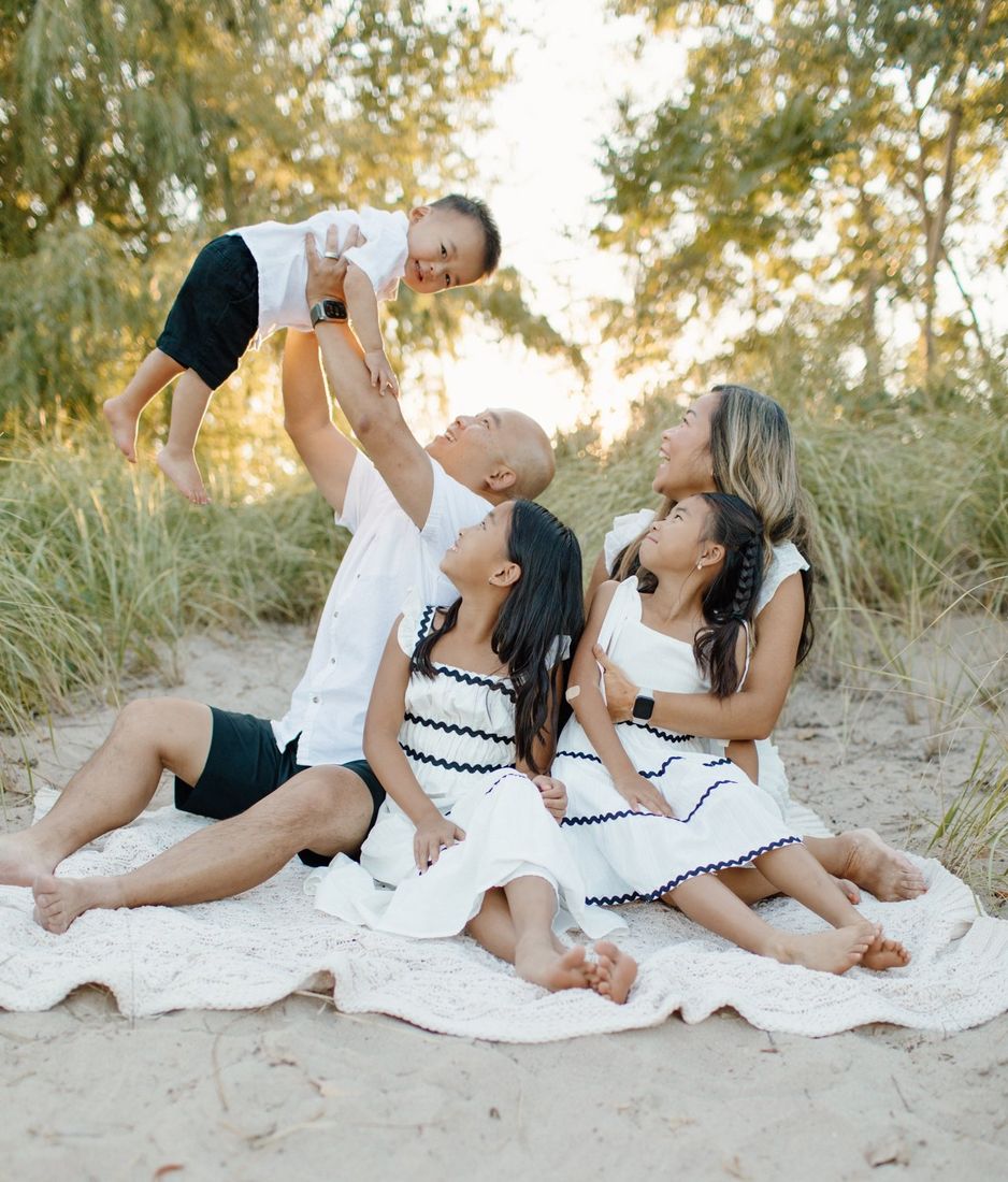 Dad lifting his son in the air as the family gathers on a beach blanket at golden hour — Home Team Photography