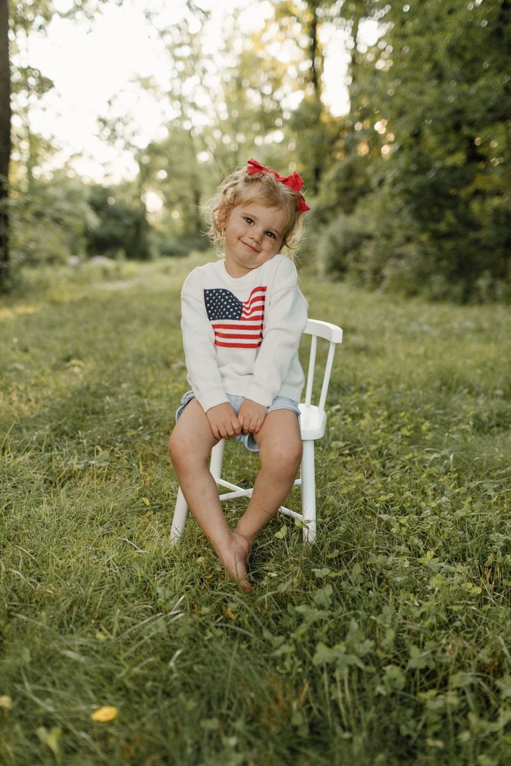 Toddler girl in a stars-and-stripes sweater sitting on a white chair in a sunlit field — Home Team Photography