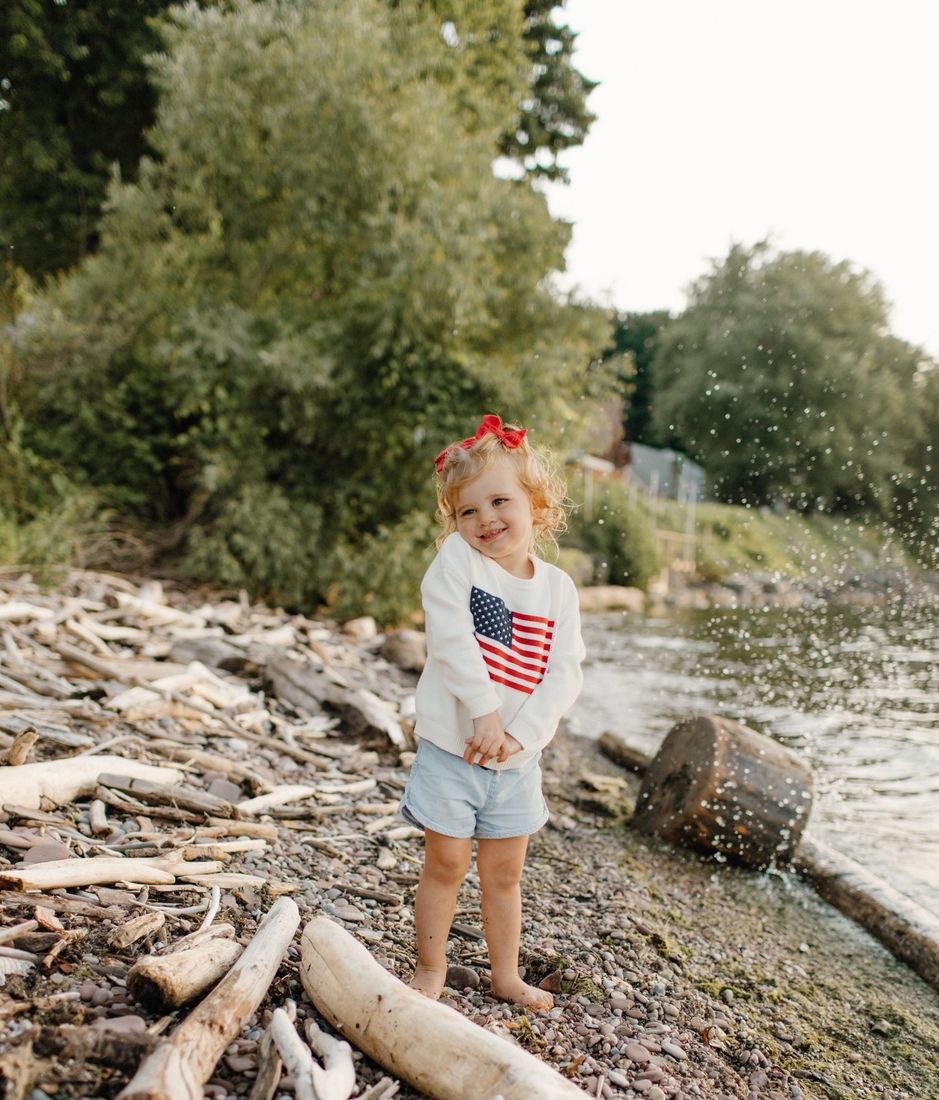 Toddler girl in a stars-and-stripes sweater by the water at golden hour — Home Team Photography