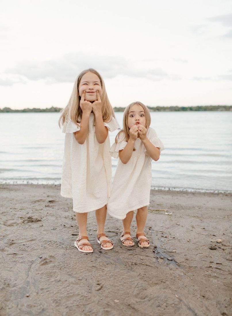 Two young sisters in cream dresses making silly faces on the lakeshore — Home Team Photography
